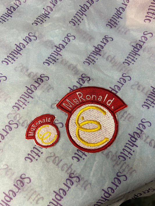 White tissue paper background, with 2 embroidered patches. Both are white with red outline, with “MgRonald.” In white lettering