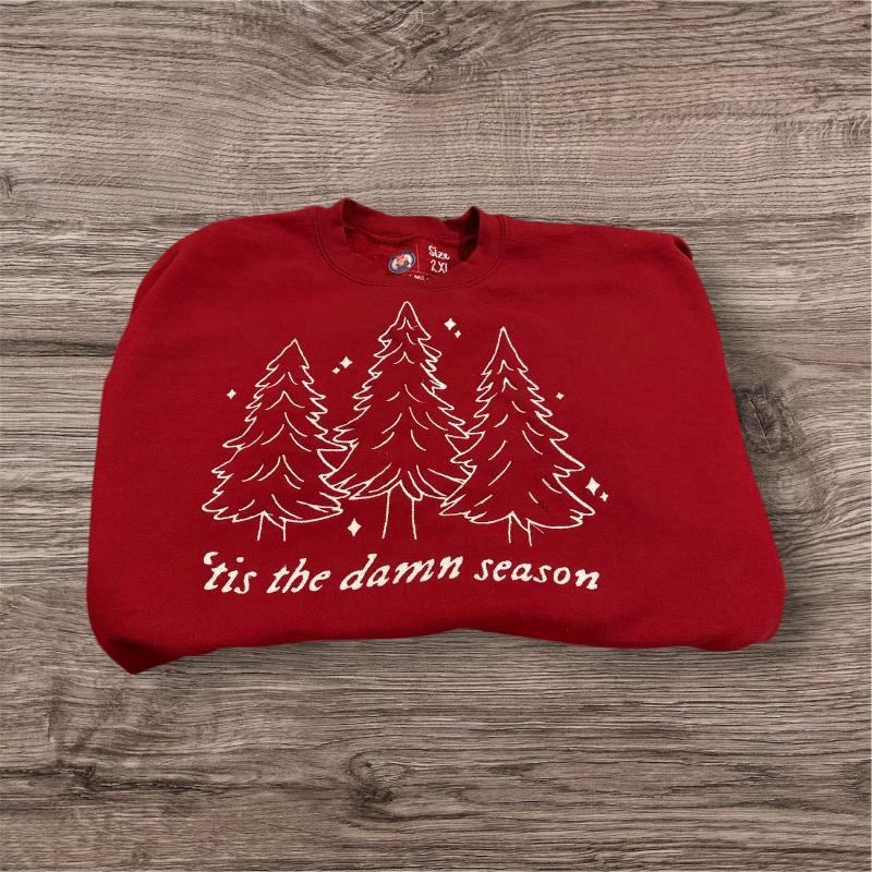 A red sweatshirt with white evergreen trees embroidered in white and " 'tis the damn season" embroidered under it also in white (shirt is brighter red in the photo than in person)