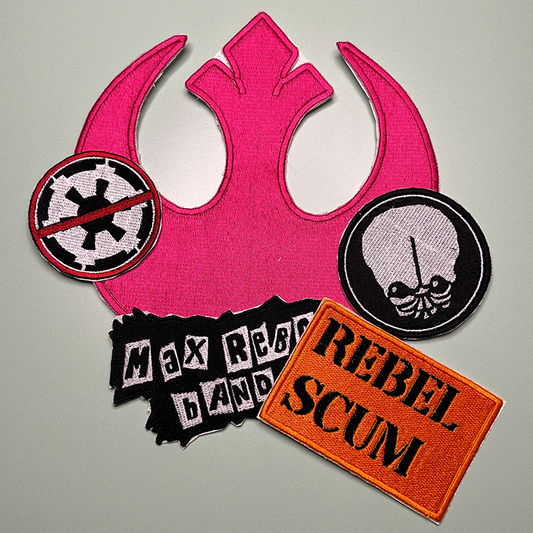 Square photo of 5 patches. The back patch is a large pink Star Wars Rebel design, with a patch of the Empire next to it and it has a crossed circle in red in front of it. On the right is a black and white patch with the likeness of Bith, in a black circle with white and black outlines. A Max Rebo Band patch with white boxes around black lettering is in the forefront, with a Rebel Scum patch in orange with black stencil style lettering in front of the Max Rebo patch