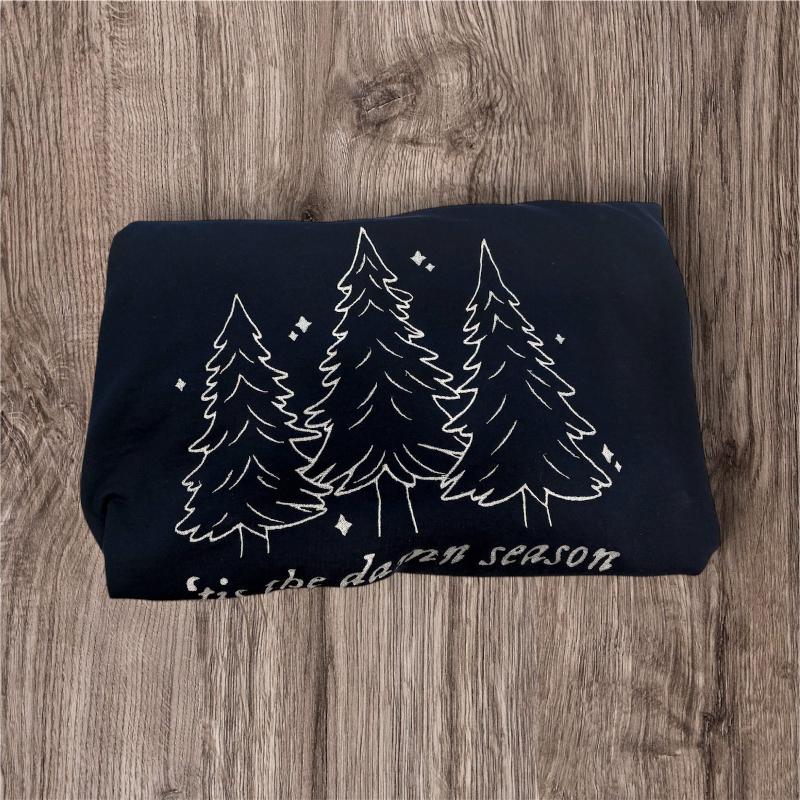 Navy blue sweatshirt, folded with white evergreen trees embroidered on it with “tis the damn season” also in white under