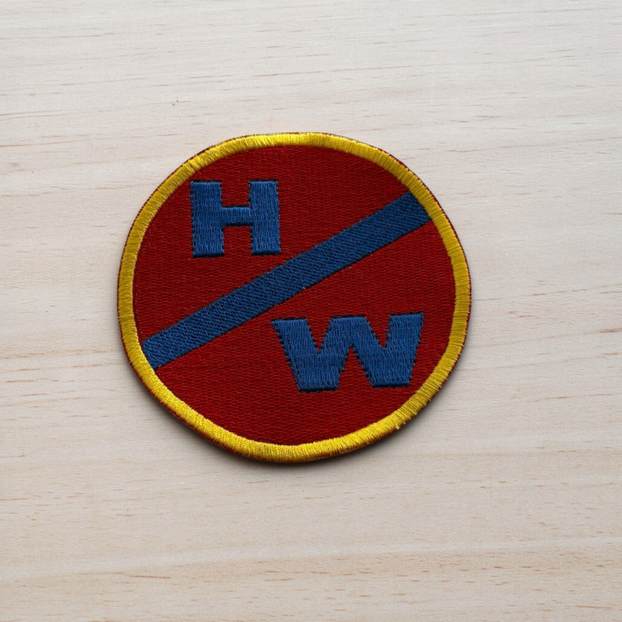 Circular patch with 'HW' letters on a red background, bordered by yellow and blue, on a light wood surface.