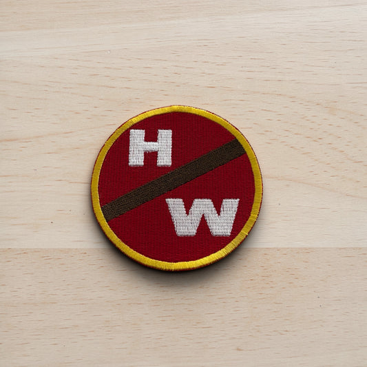 Round patch with 'H' and 'W' on a red background, bordered by yellow and brown, on a wooden surface.