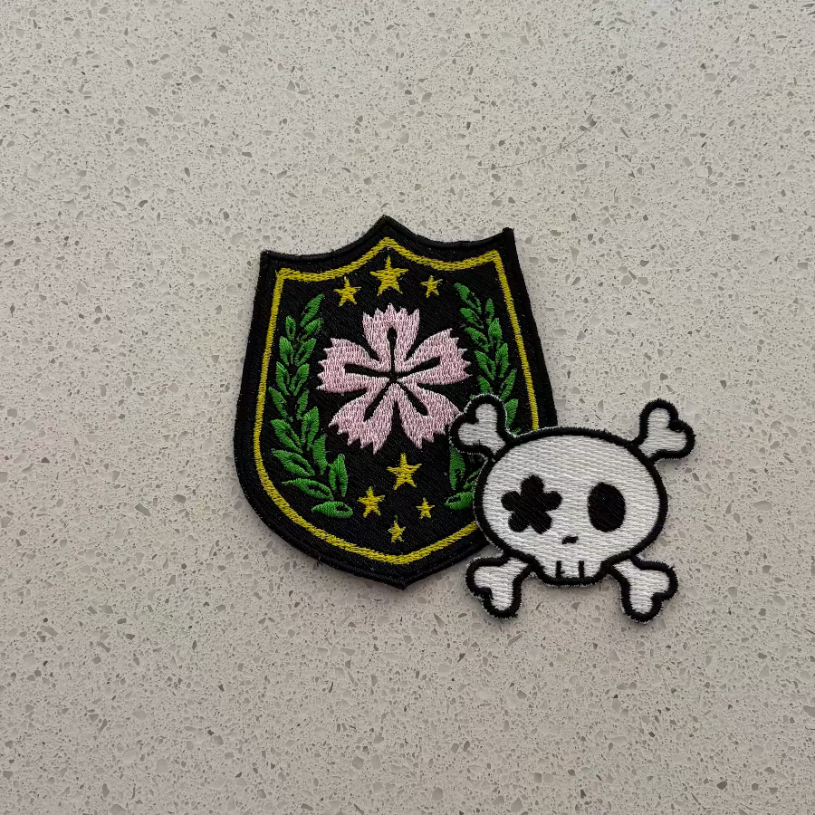 Two embroidered patches - one with a shield design and a pink flower in the center, with 7 stars in yellow with green sprigs on either side. Overlapping it is a skull and crossbones with one eye socket in the shape of a flower