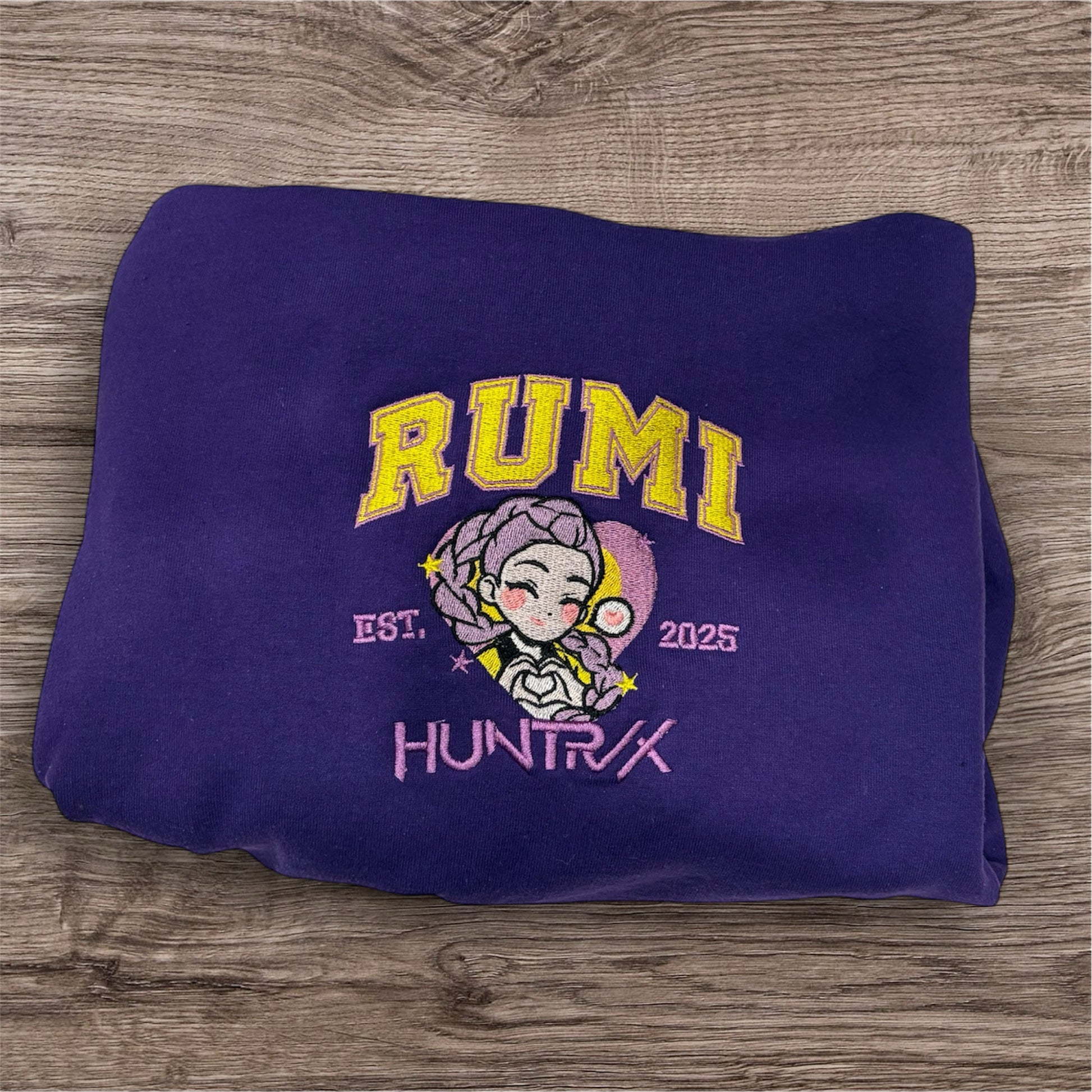 A purple sweatshirt, on top of a light woodgrain backdrop. The shirt is embroidered with “RUMI” in yellow with a light purple border, with a portrait of Rumi from KPop Demon Hunters under it, with “EST 2025” around her and “HUNTRIX” under that in lavender.