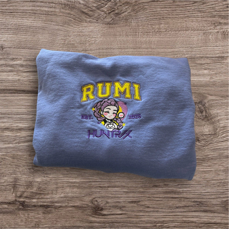A light purple sweatshirt, on top of a light woodgrain backdrop. The shirt is embroidered with “RUMI” in yellow with a light purple border, with a portrait of Rumi from KPop Demon Hunters under it, with “EST 2025” around her and “HUNTRIX” under that in lavender