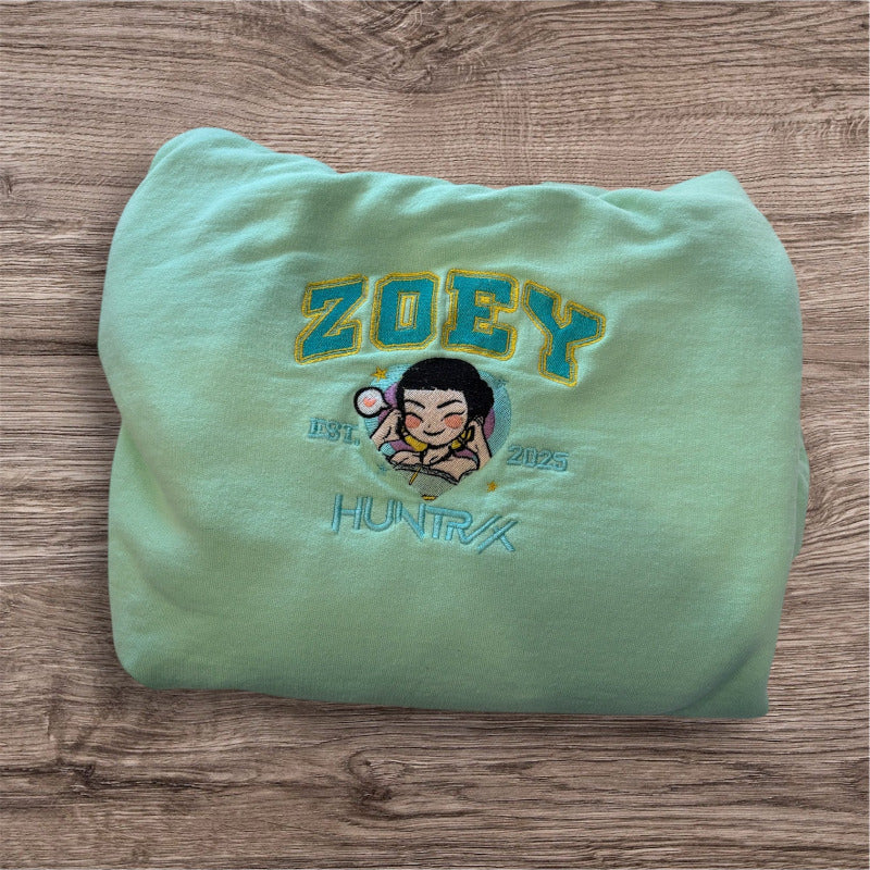 A mint green sweatshirt, on top of a light woodgrain backdrop. The shirt is embroidered with “ZOEY” in greenwith a yellow border, with a portrait of Zoey from KPop Demon Hunters under it, with “EST 2025” around her and “HUNTRIX” under that in green.