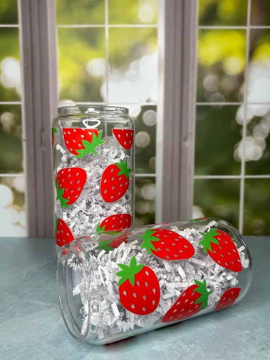Two clear glass tumblers, both with decals of strawberries covering them. One is standing up, the other is on its side. Both are filled with crinkled white paper.