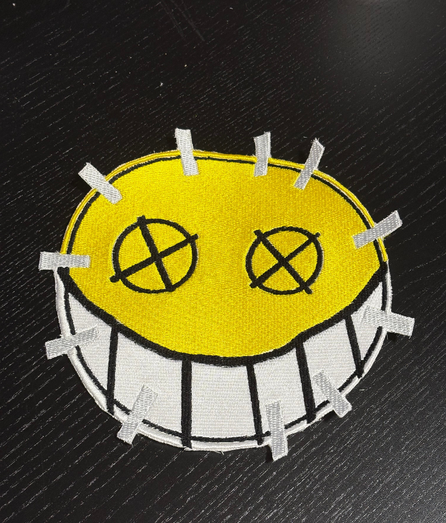 A yellow smiley face embroidered patch, with a row of teeth along the bottom. The eyes are circles outlined in black, with an “X” over each