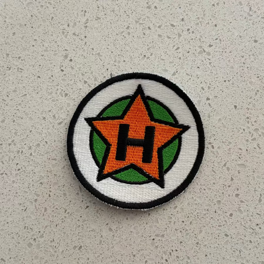 A circular embroidered patch, with black outlines with white and green backgrounds, and an orange star in the center with a black “H”