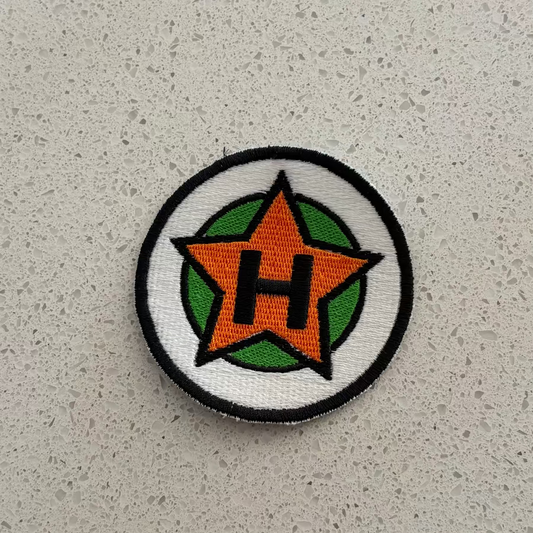 A circular embroidered patch, with black outlines with white and green backgrounds, and an orange star in the center with a black “H”