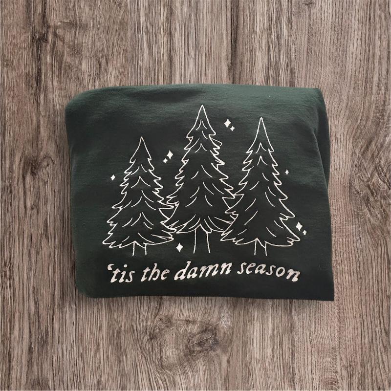 Forest green sweatshirt folded on a wood grain background. In white is embroidered 3 evergreen trees and "tis the damn season" embroidered under it