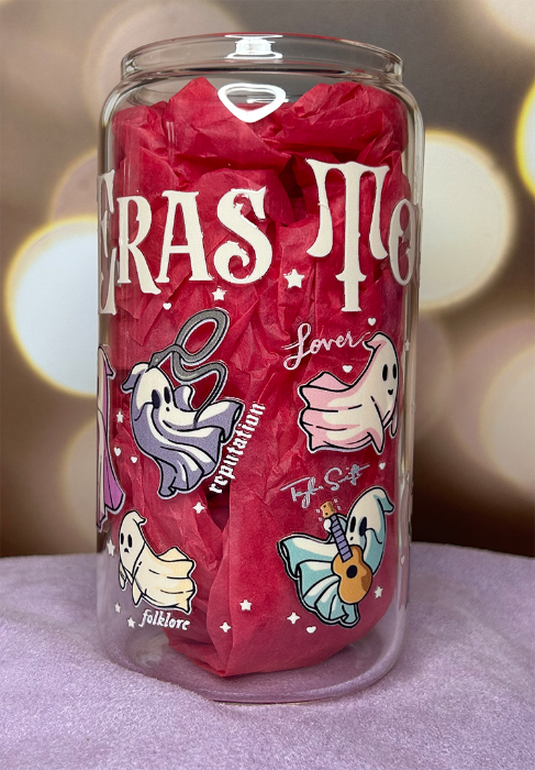 Clear glass can tumbler with "The Eras Tour" on it, with sheet style ghosts representing Taylor Swift albums on it, and the titles of each album. The cup is stuffed with pink tissue paper to make the decals more visible