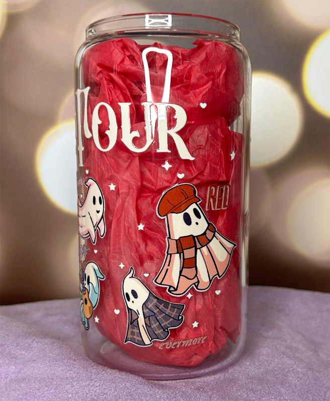 Clear glass can tumbler with "The Eras Tour" on it, with sheet style ghosts representing Taylor Swift albums on it, and the titles of each album. The cup is stuffed with pink tissue paper to make the decals more visible