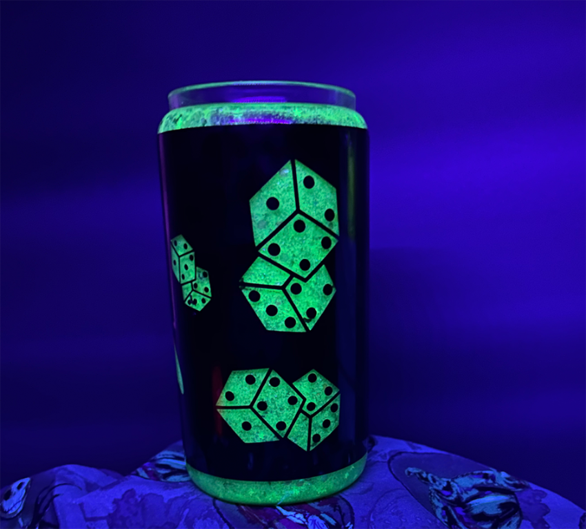 Glitter globe glass tumbler, with neon green glow in the dark glitter - under a black light, so the glitter is glowing in the dark. There is black vinyl wrapped around it, with dice cut out of the vinyl design.