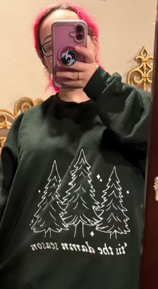 Forest green sweatshirt, oversized on the model. In white is embroidered 3 evergreen trees and "tis the damn season" embroidered under it